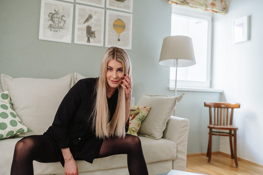 Nina Milenkovics sitzt auf einer beigen Couch. Sie hat ein schwarzes Kleid und eine schwarze Strumpfhose an und sieht direkt in die Kamera. 