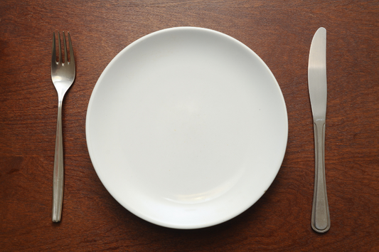 Ein weißer, leerer Teller steht auf einem braunen Tisch. links eine silberne Gabel, rechts ein silbernes Messer.