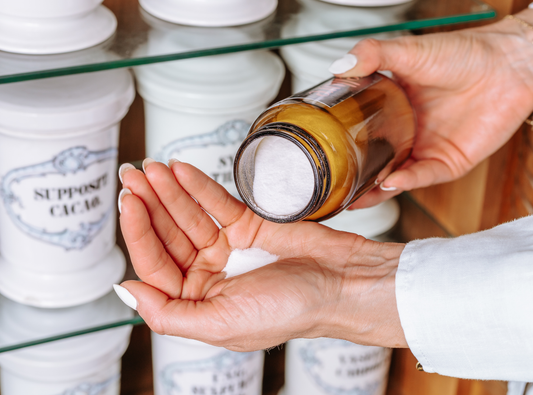 Eine Frau steht vor einem Regal mit alten Apothekengefäßen und hat in der rechten Hand ein braunes Glas mit einem weißen Pulver. Dieses schüttet sie sich selbst in ihre linke Hand. 
