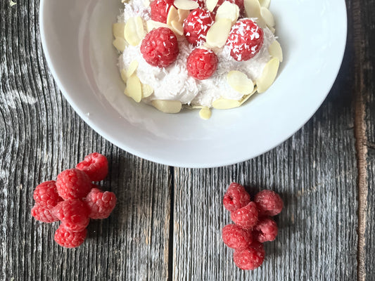 Eine weiße Schüssel mit Joghurt, Kokosraspeln und Mandelstückchen steht auf einem alten Holzbrett. Im Vordergrund liegen einige Himbeeren. 