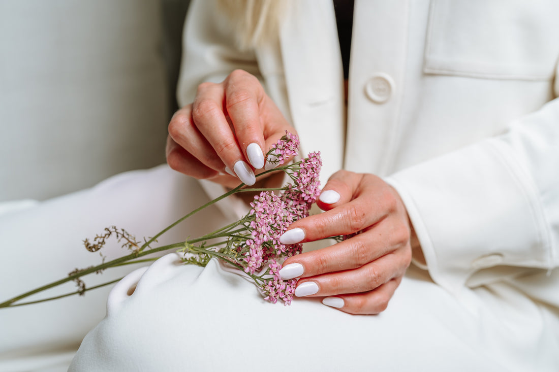 Man sieht die Hände einer Frau mit schön lackierten, weißen Fingernägeln, die eine rosarote Blume in der Hand halten. 