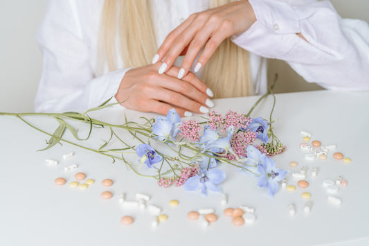 Man sieht ganz viele verschiedene Pillen auf einem weißem Tisch liegen und zusätzlich 2 verschiedene Blumen. Im Hintergrund sieht man eine Frau mit blonden Haaren und einer weißen Bluse sitzen.