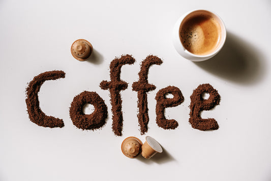 Im Bild wurde "Coffee" mit Kaffeepulver auf einen weißen Tisch geschrieben. Außerdem liegen 3 Kaffeekapseln herum und eine Tasse mit schwarzem Kaffee steht in der linken oberen Ecke.