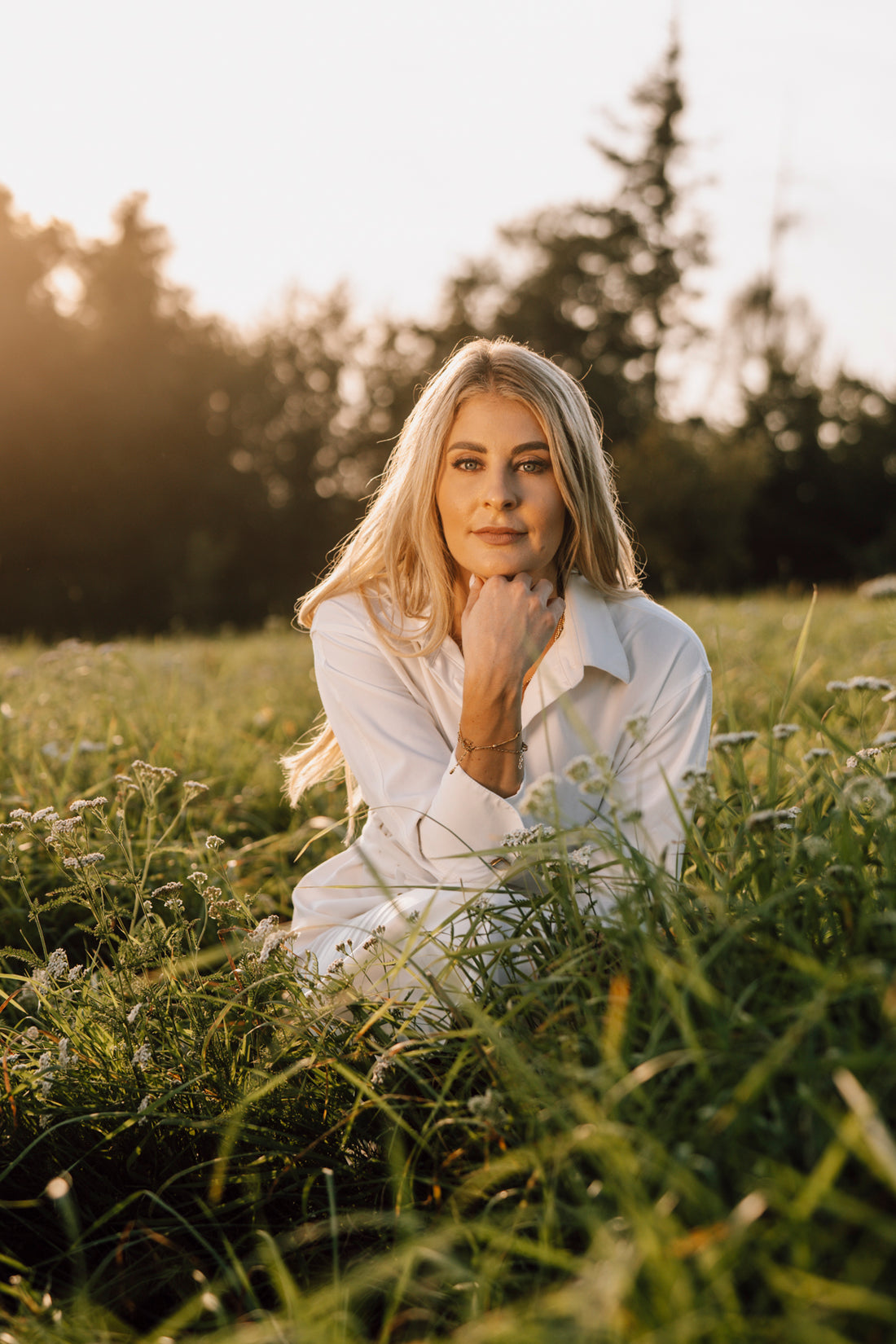 Nina Milenkovics hockt im Gras und schaut direkt in die Kamera. Sie hat offene Haare und trägt ein weißes Sommerkleid.