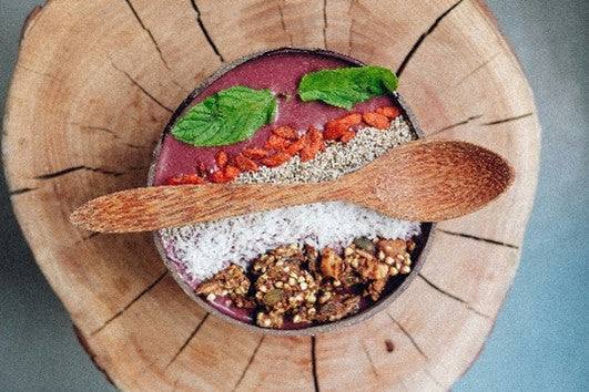 Auf einer Holzscheibe steht eine Schale mit einer Bowl gefüllt mit verschiedenen Lebensmitteln. Darauf liegt ein großer Holzlöffel.