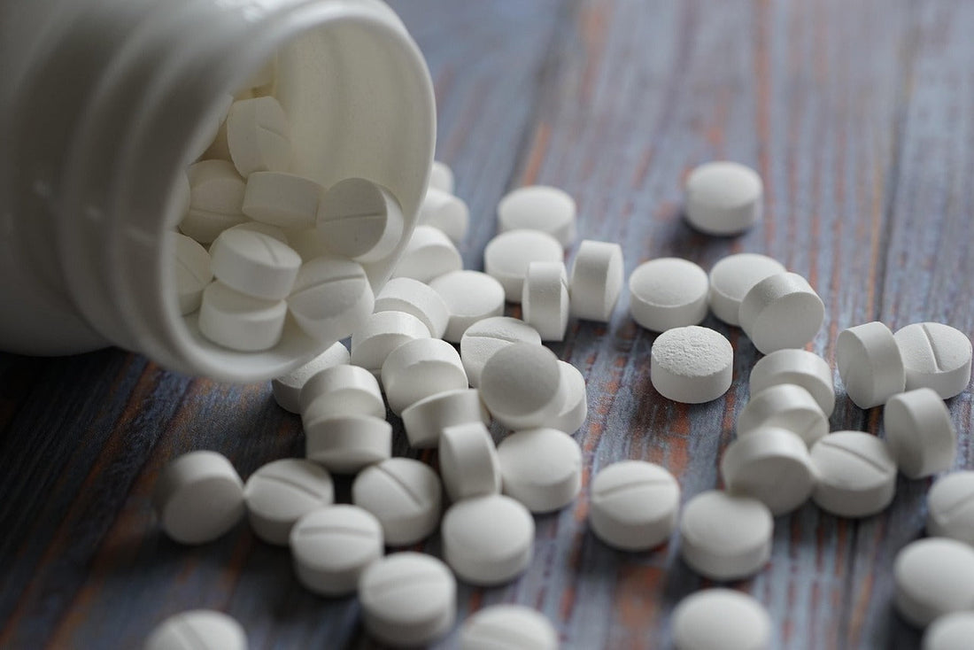 Auf einem Holztisch liegt eine geöfnette Tablettendose und viele weiße Tabletten sind überall verteilt.