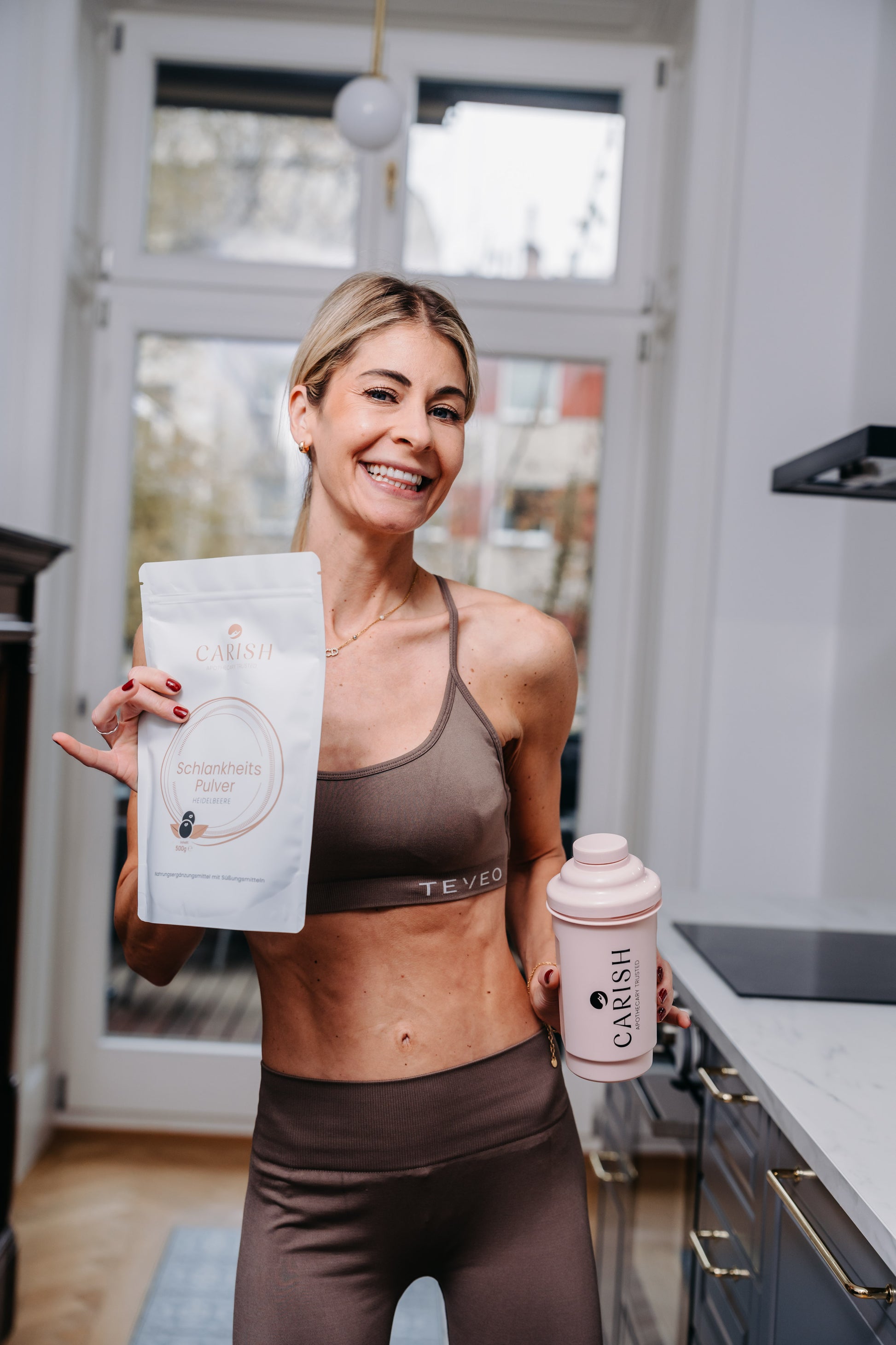 Die Apothekerin Nina Milenkovics steht in der Küche und hat in der rechten Hand das Schlankheitspulver Heidelbeere und hält in der linken Hand einen rosaroten Shaker. 