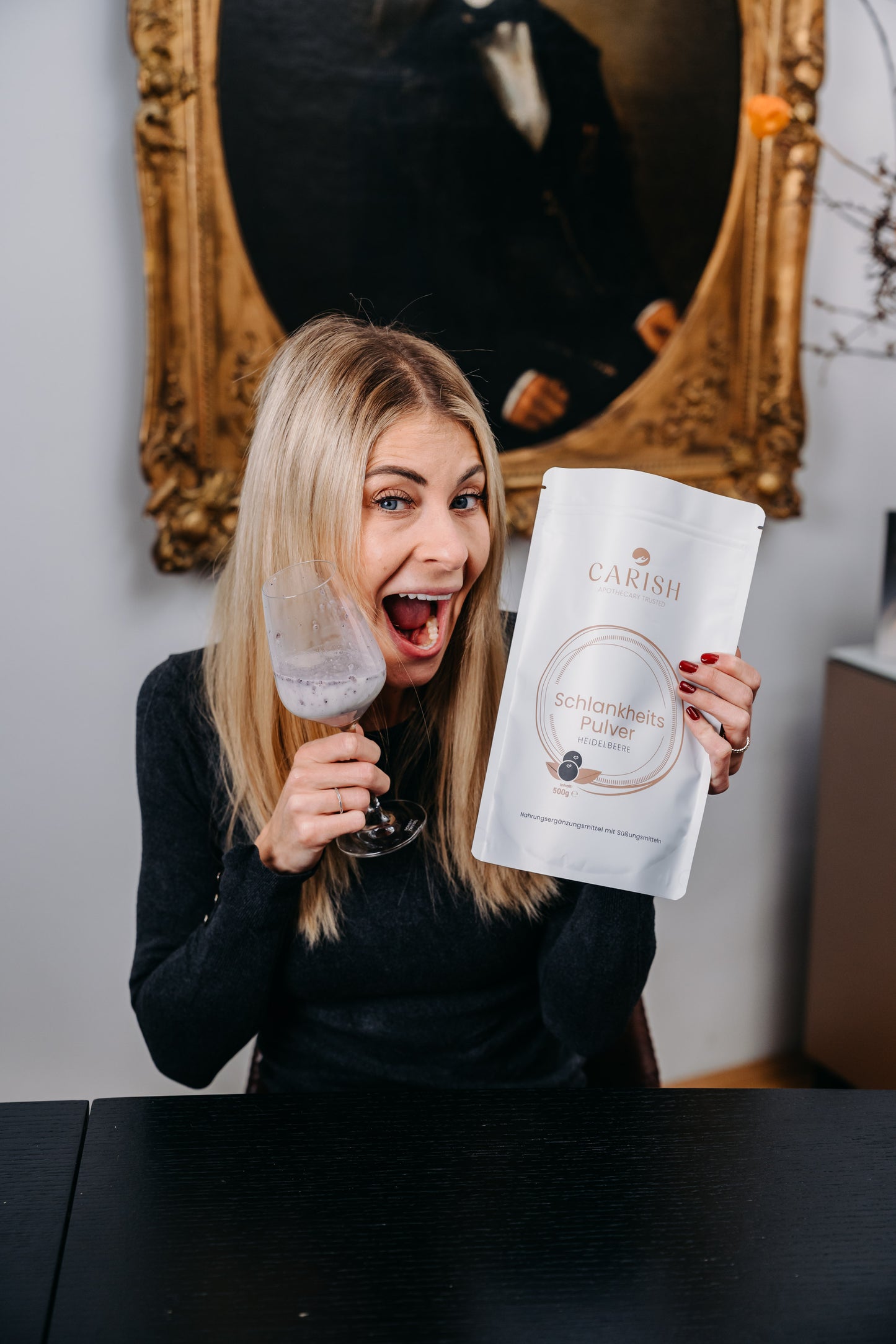 Die Apothekerin Nina Milenkovics sitzt an einem Tisch und hält das Schlankheitspulver Heidelbeere und ein Glas mit dem angerührten Pulver in ihren Händen. 