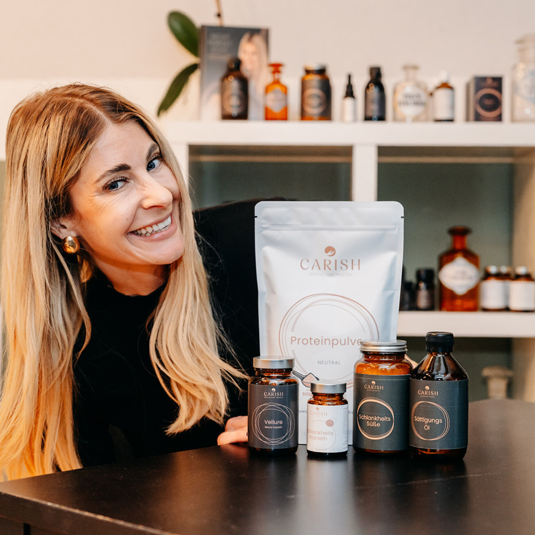 Die Apothekerin Nina Milenkovics sitzt an einem Tisch, lächelt in die Kamera und hat vor sich die 5 Produkte des Daily must haves Paketes von Carish stehen. 