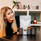 Die Apothekerin Nina Milenkovics sitzt an einem Tisch, lächelt in die Kamera und hat vor sich die 5 Produkte des Daily must haves Paketes von Carish stehen. 