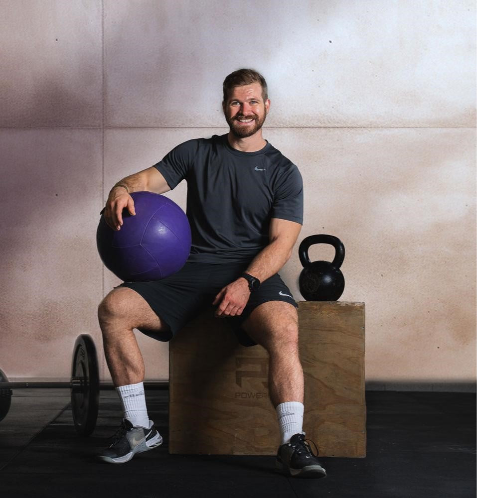 Der Fitnesstrainer Daniel Sommer sitzt auf einer Kiste und hat einen blauen Gymnastikball in der Hand. 