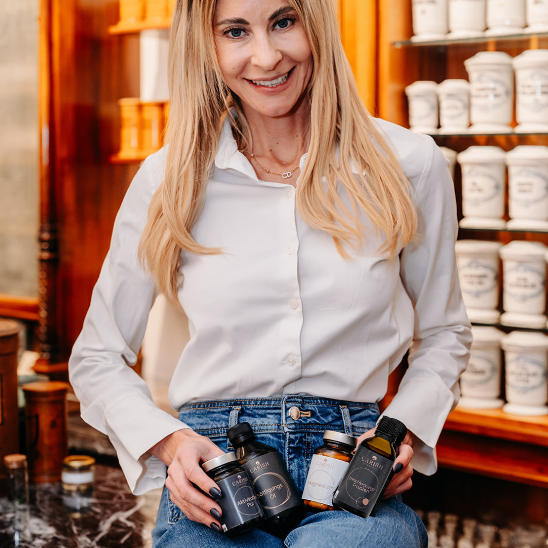 Nina Milenkovics sitzt auf einem Tisch, im Hintergrund alte Apothekengefäße und hält die 4 Produkte des Detoxpaket von Carish in ihren Händen. 
