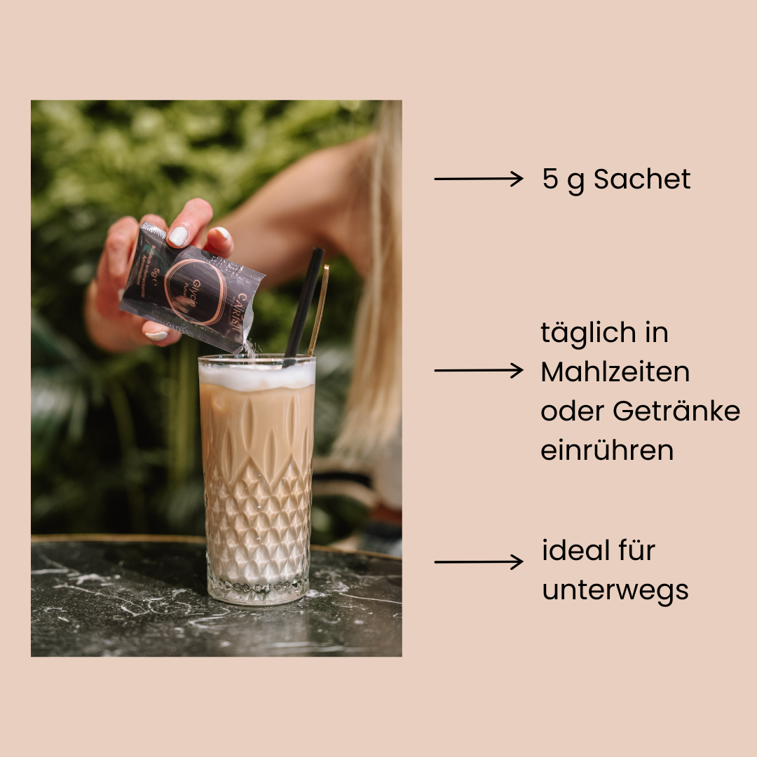 Eine Frau sitzt bei einem Tisch und hat einen Latte Macchiato vor sich stehen. Mit der rechten Hand schüttet sie von einem geöffneten Glycin Sachet das Pulver in den Kaffee. 