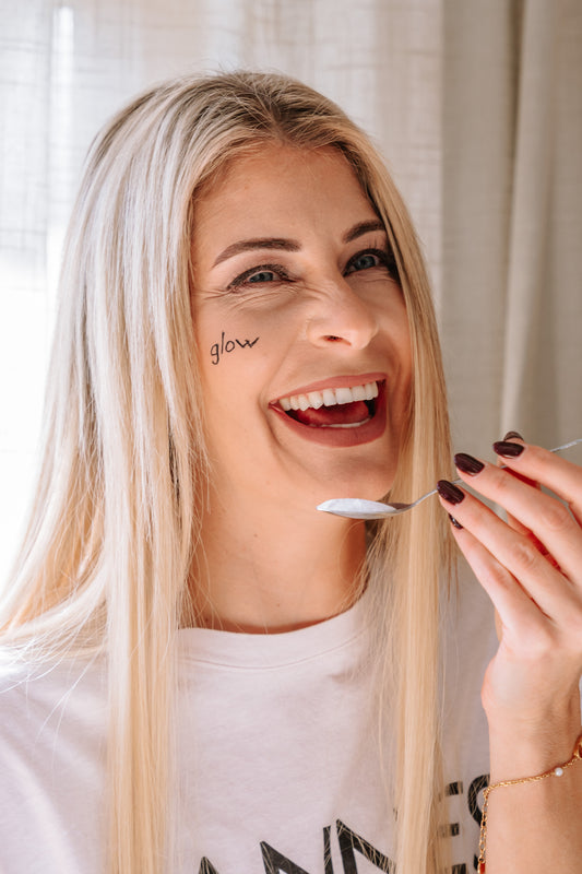Nina Milenkovics lacht in die Kamera, hat einen Löffel mit Glycin in der Hand und das Wort "glow" auf die Wange geschrieben. 