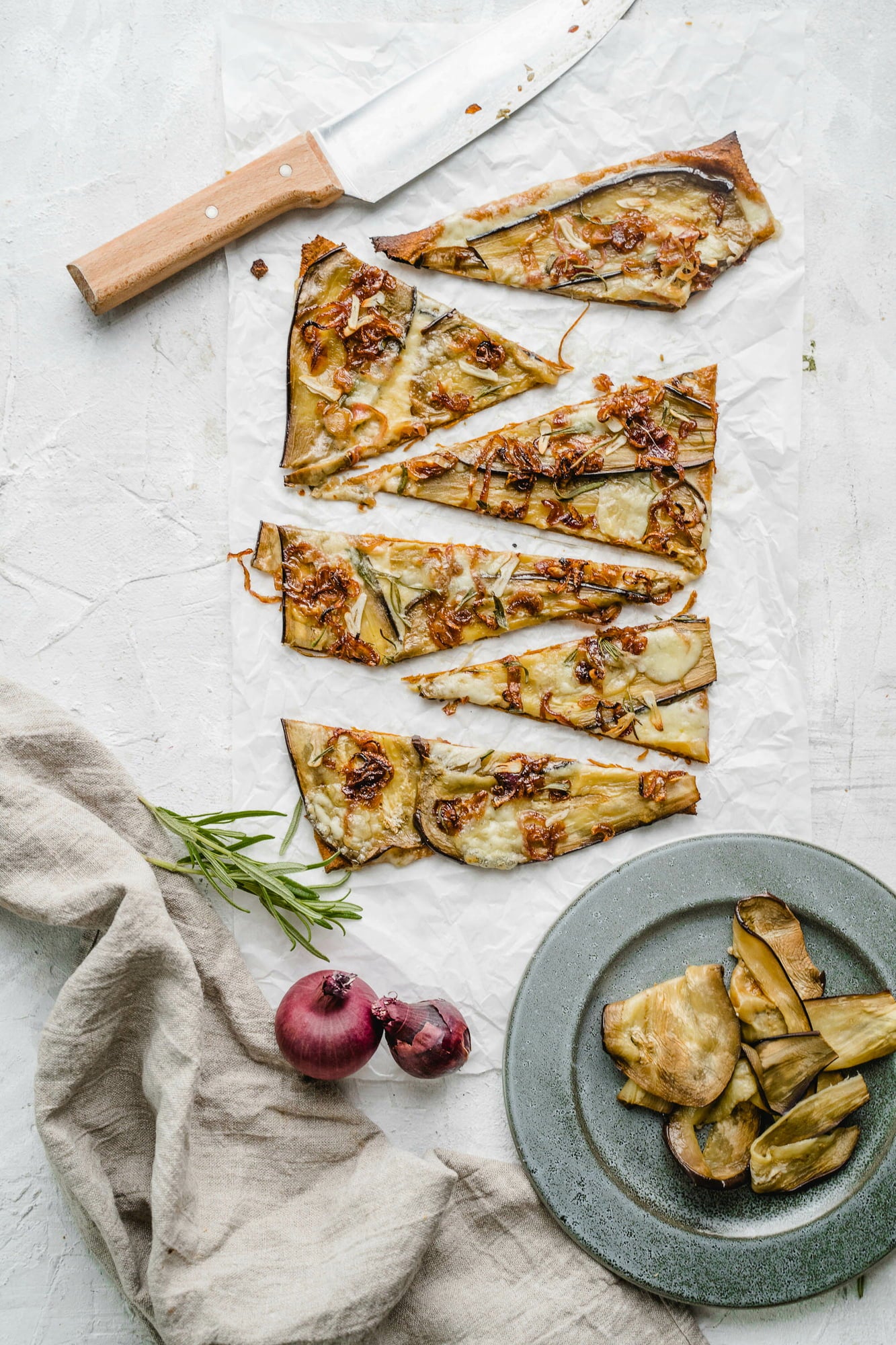 Serviervorschlag eines belegten veganen Pizzabodens von oben fotografiert. Aufgeschnittene Pizza, ein Messer, Rosmarin, Zwiebel und ein Teller mit Pizzastücken ist zu sehen.